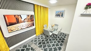 Habitación moderna con televisor de pantalla plana montado en la pared, una mesa de comedor con sillas, una cortina amarilla y una fotografía de un paisaje urbano.