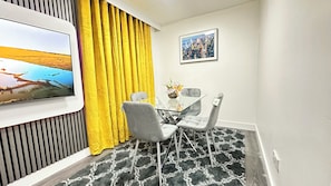 Un espace de salle à manger moderne avec une table en verre, des chaises grises, un rideau jaune et un tableau représentant un paysage urbain accroché au mur.