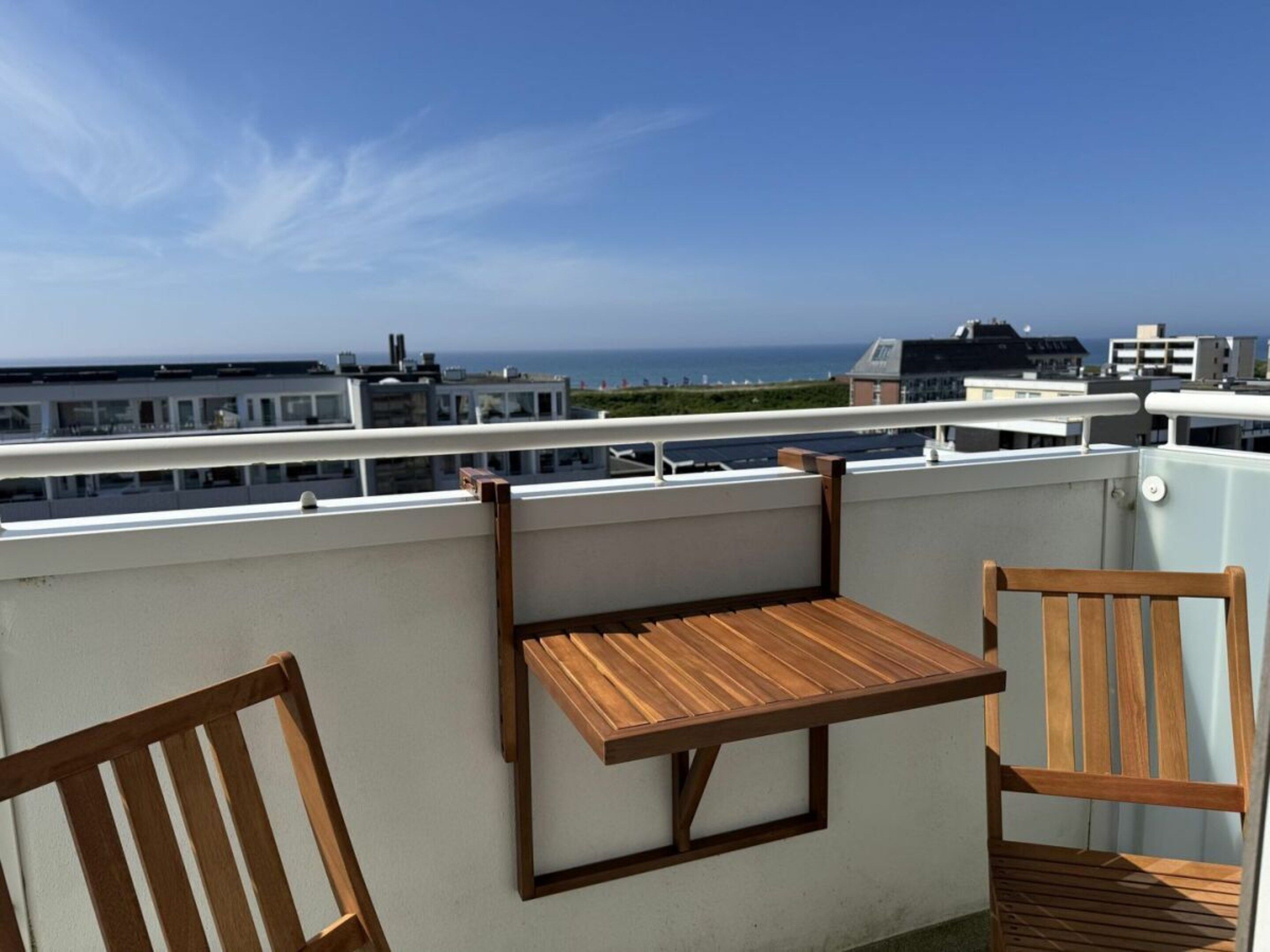 Een balkon met een houten tafel en twee stoelen, met uitzicht op de kust en gebouwen in de verte.