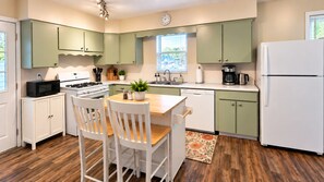 A modern kitchen with white cabinets, a microwave, stove, and a refrigerator. There is a dining table with chairs and a window with blinds. - Comfortable Home Base | Near ND, IUSB & Hospitals (Mishawaka)