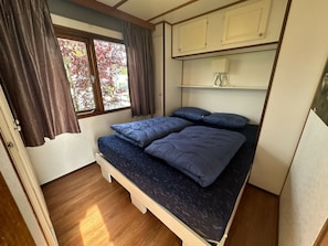 A compact bedroom with a single bed, a window with curtains, and a small shelf with a lamp.