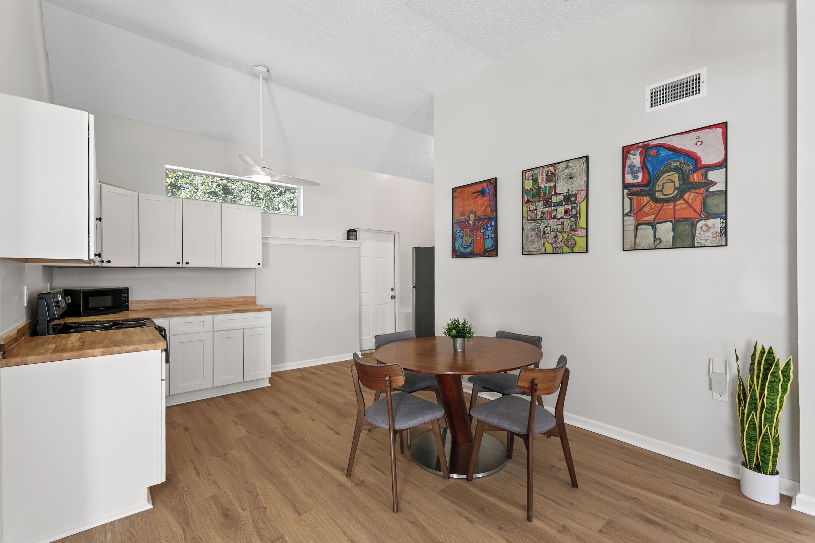 A modern kitchen with white cabinets, a microwave, and a dining area with a round table and chairs. There are three abstract paintings on the wall.