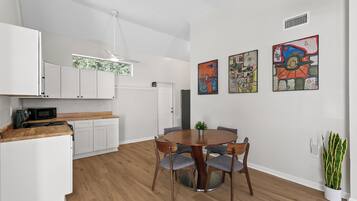A modern kitchen with white cabinets, a microwave, and a dining area with a round table and chairs. There are three abstract paintings on the wall.