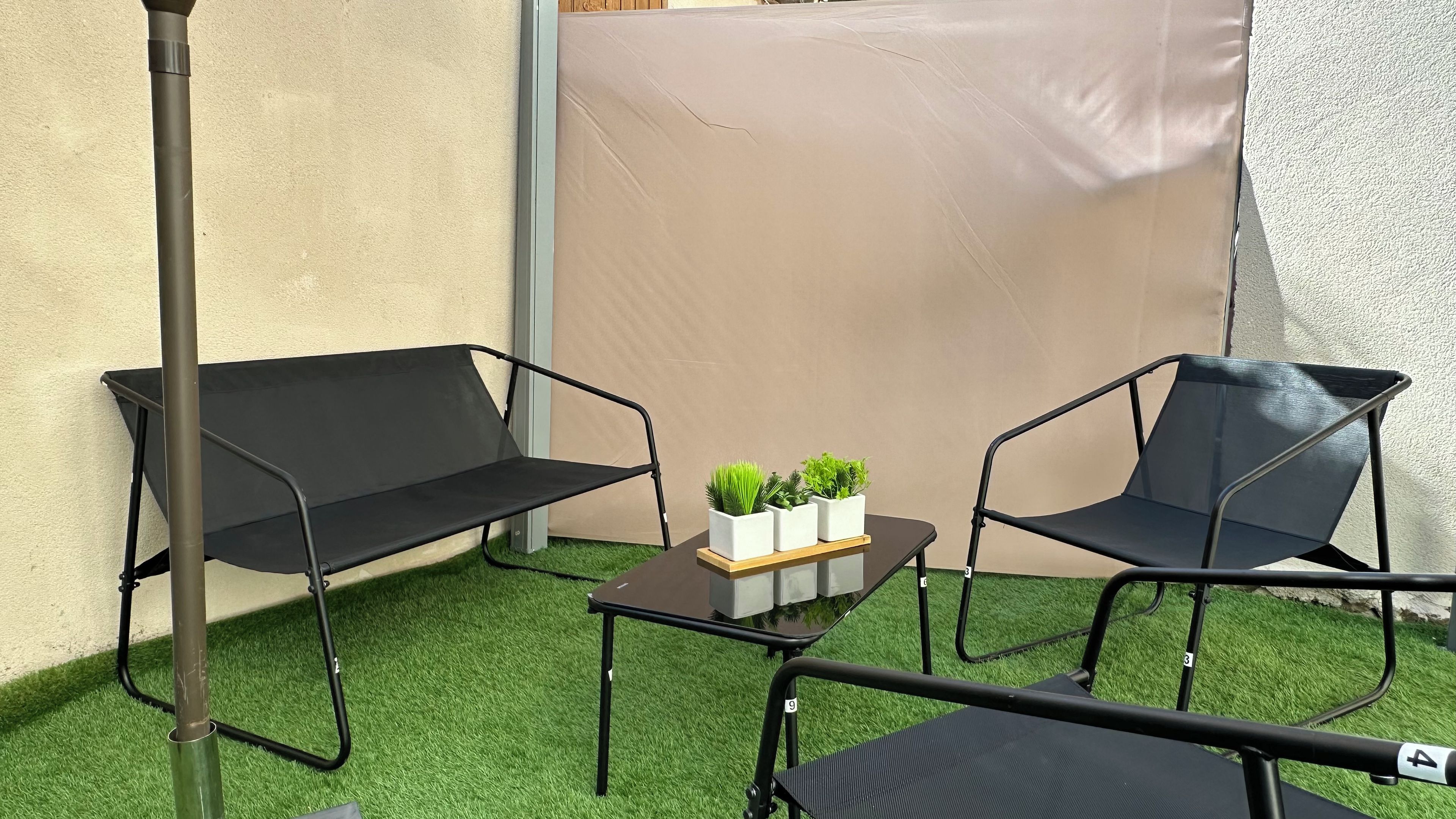Un petit espace extérieur aménagé avec deux chaises en métal noir et une table, entouré de gazon synthétique et d’un mur de couleur beige.