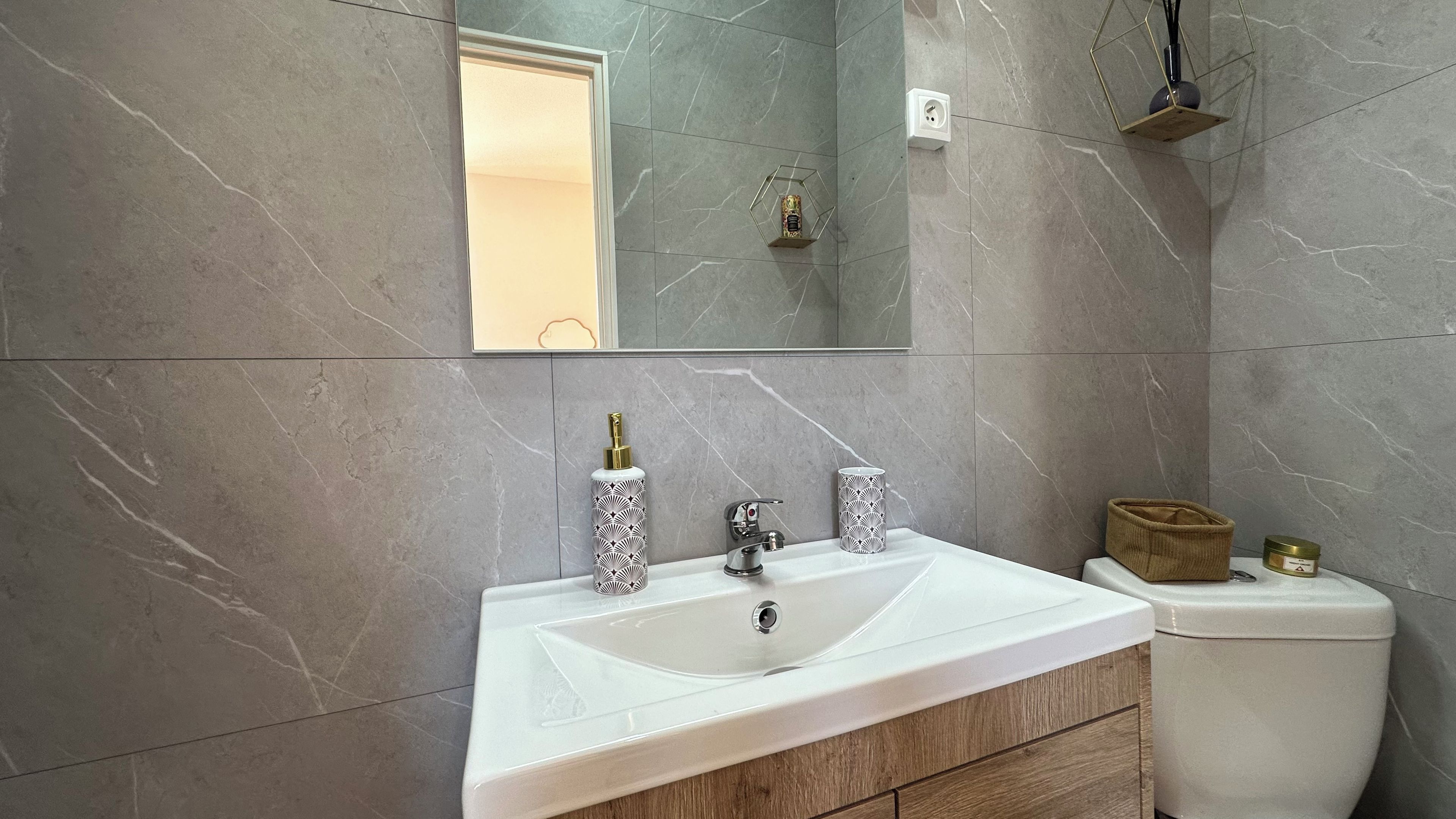 A bathroom with a marble wall, a sink with two faucets, a mirror, and a toilet.