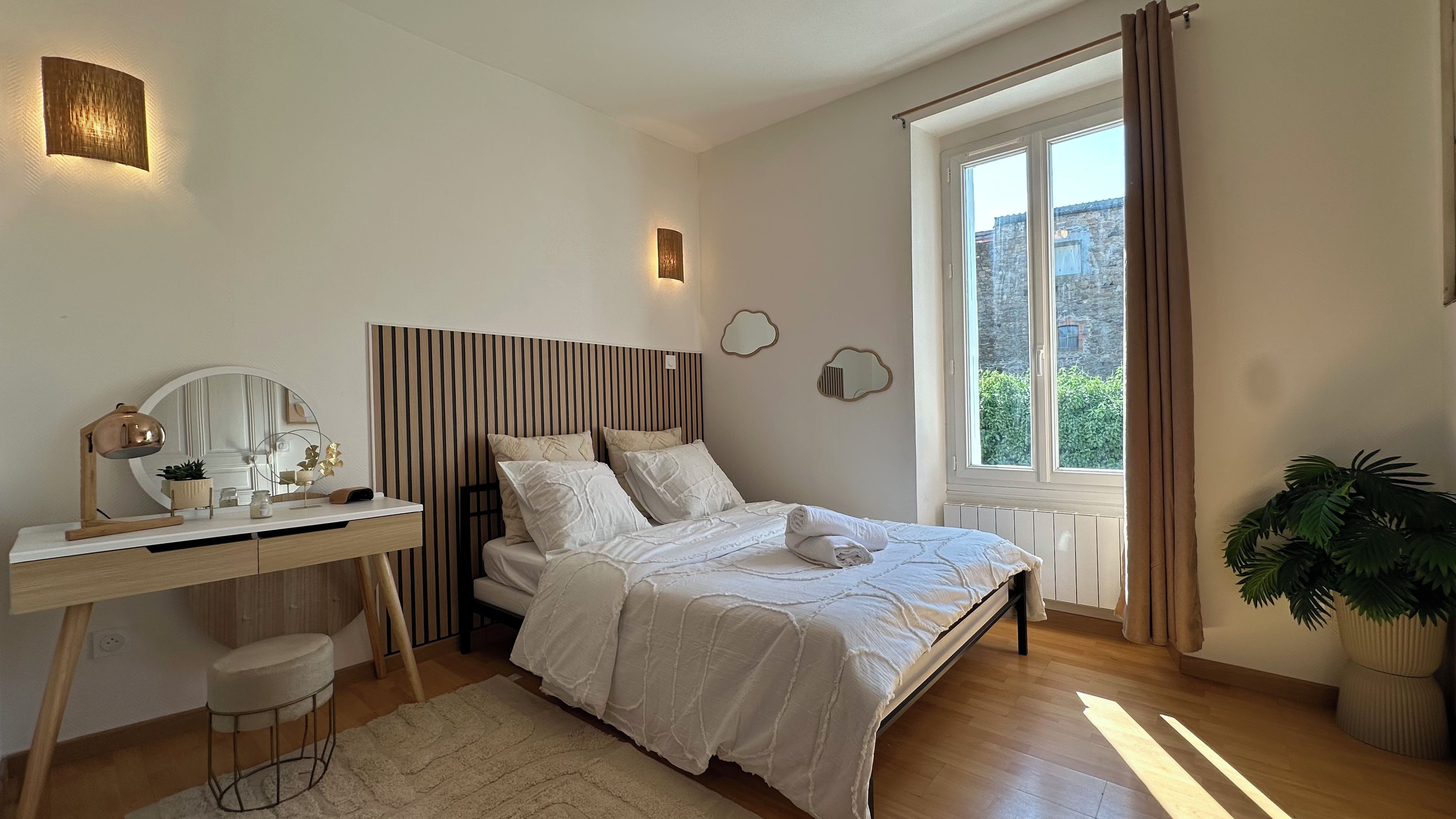 Une chambre à coucher avec un lit, une table de chevet, une lampe, un miroir et une fenêtre avec des rideaux.