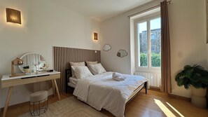 Une chambre à coucher avec un lit, une table de chevet, une lampe, un miroir et une fenêtre avec des rideaux.