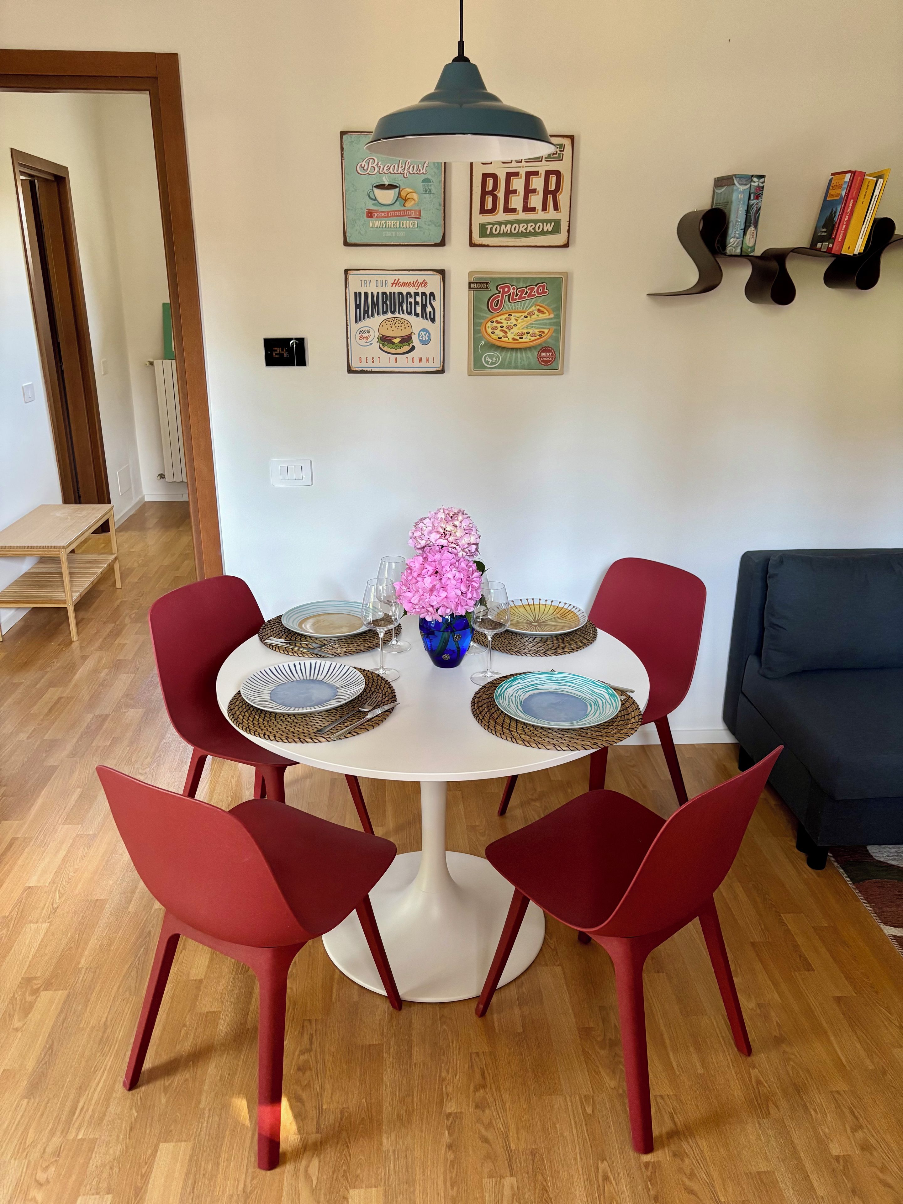A dining area with a round table, red chairs, a vase with pink flowers, and wall decor including signs for breakfast, beer, hamburgers, and pizza.