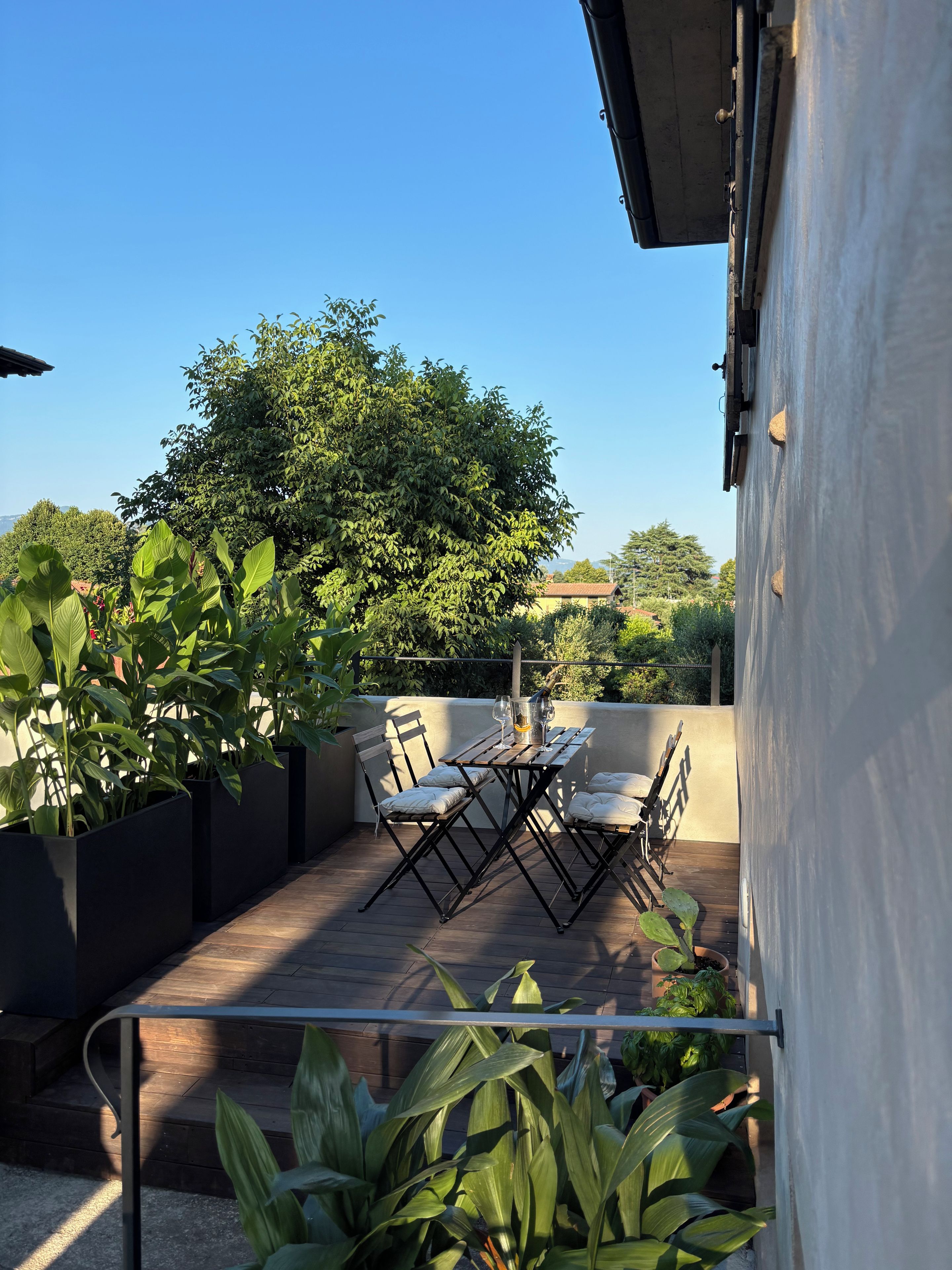 Una terrazza con tavolo e sedie, circondata da piante in vaso e vista su alberi.