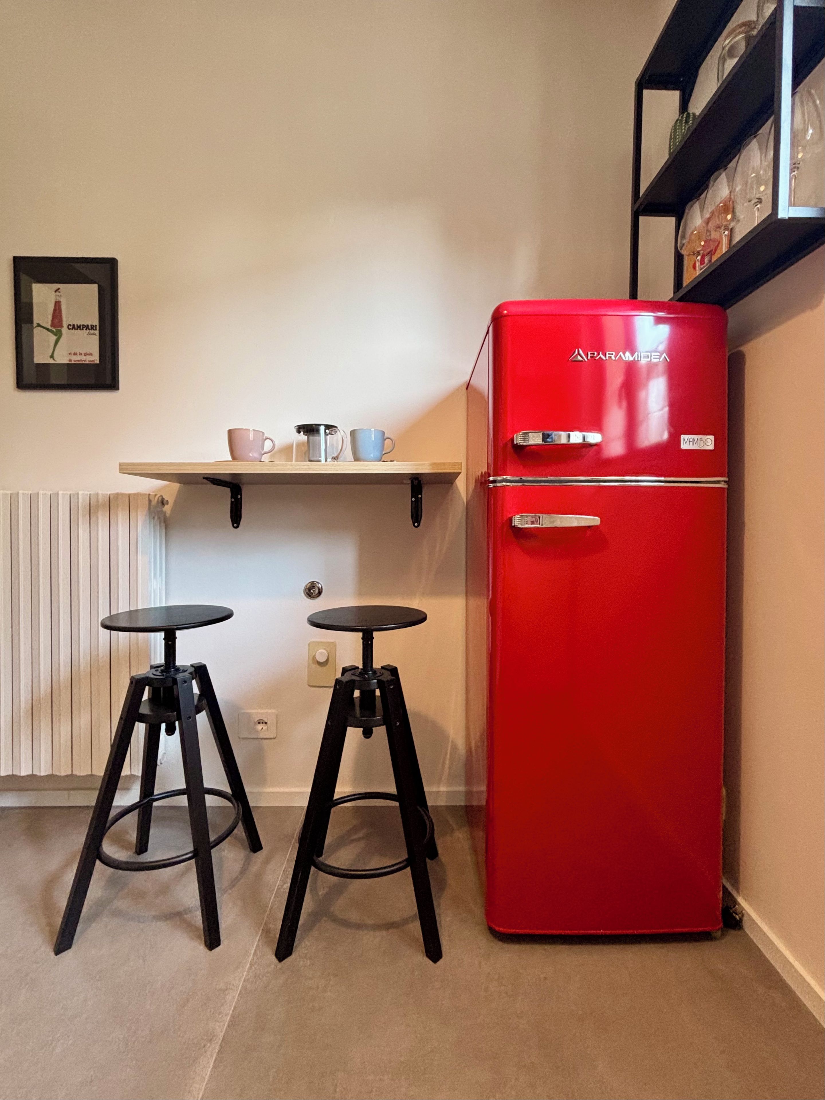 Una kitchenette compacta con un refrigerador rojo, dos taburetes negros, un estante con tazas y un cuadro en la pared.