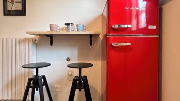 Una kitchenette compacta con un refrigerador rojo, dos taburetes negros, un estante con tazas y un cuadro en la pared.