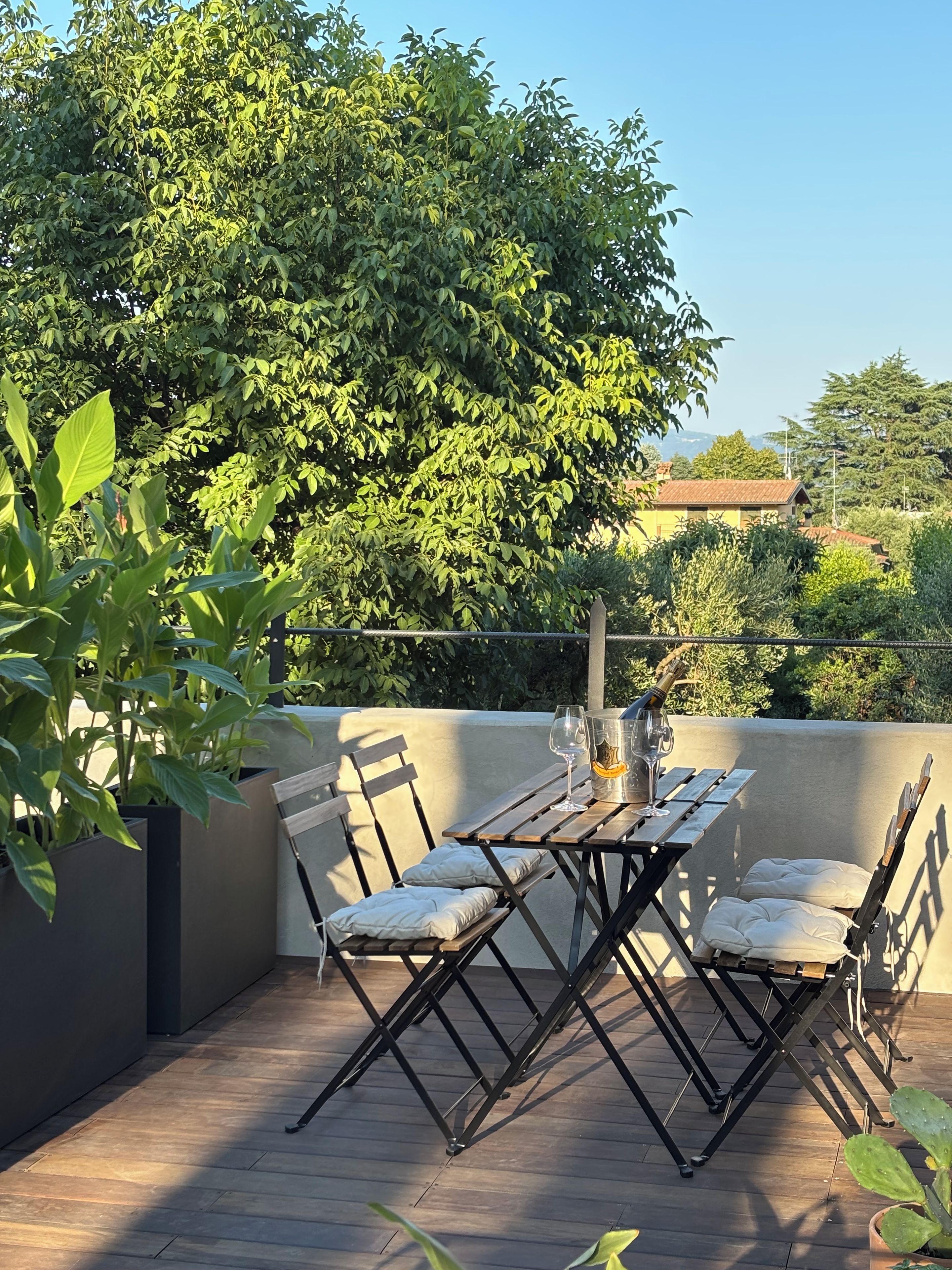 An outdoor patio with a wooden table and chairs, surrounded by greenery and a view of the sky.