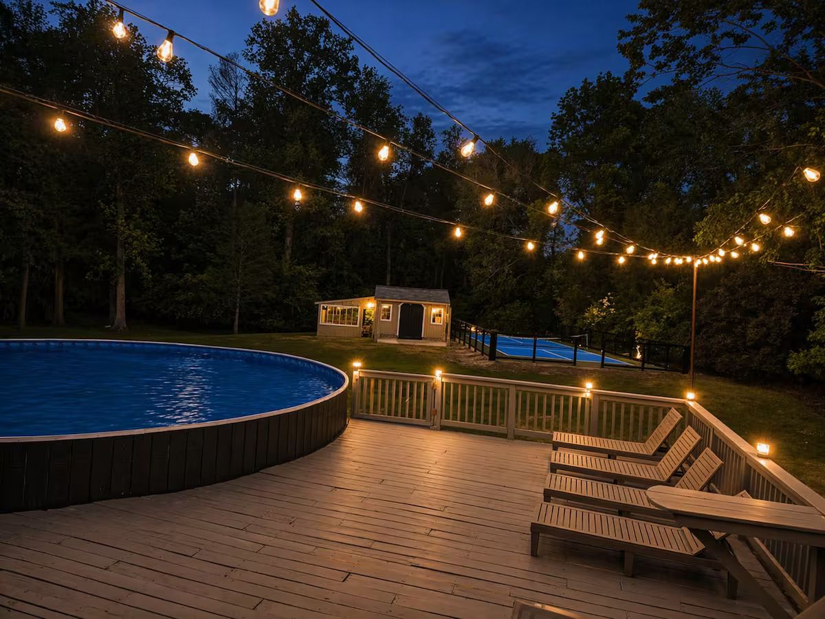 Una piscina con muelle de madera, sillas de descanso y luces decorativas.