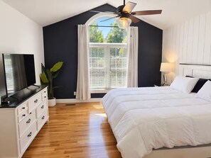 Un dormitorio con cama, televisor, ventilador de techo y ventana con cortinas.