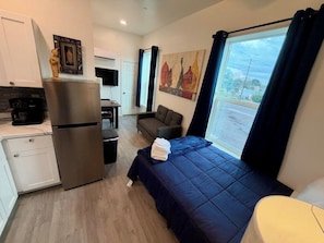 Quarto de hotel compacto com cama, pequeno espaço de cozinha, área de estar e janela com cortinas.