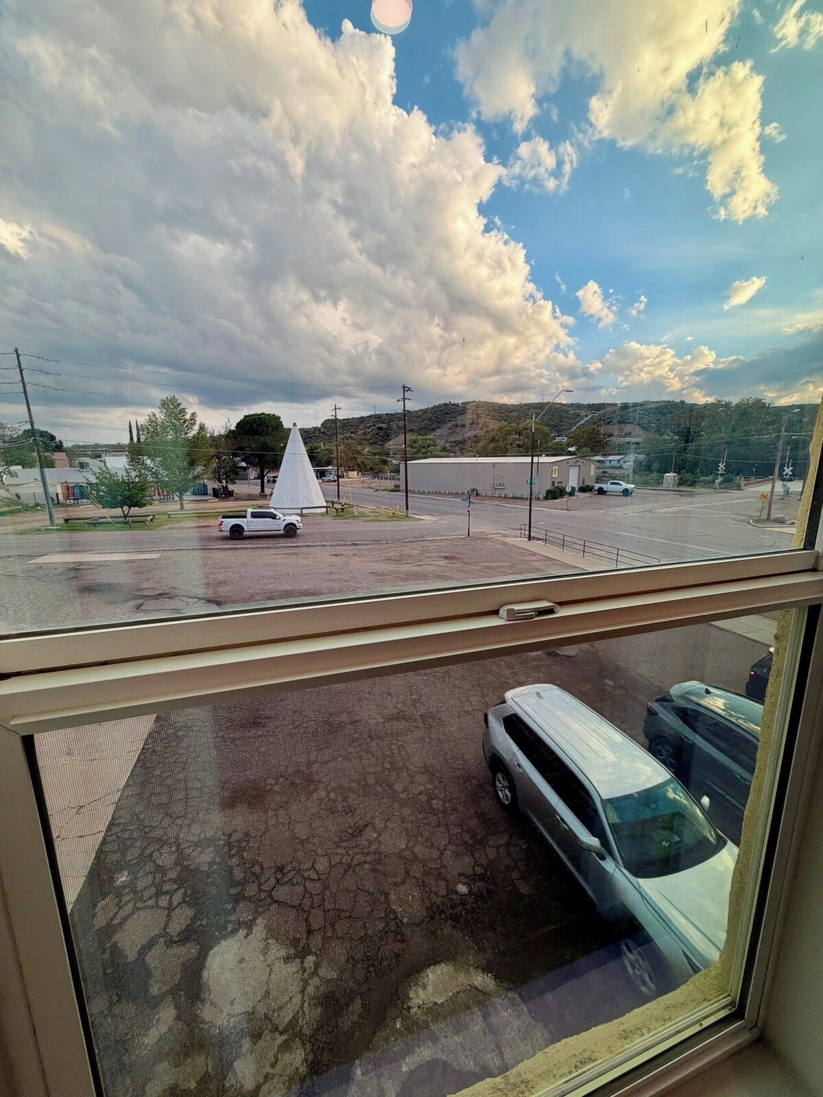 A view from a window overlooking a parking area with a white car and a white van, a traffic cone, and a building with a flat roof in the distance.