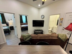 Espaço compacto com sofá, TV de tela plana, mesa de jantar e área de cozinha ao fundo.