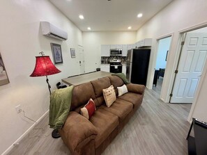 Uma sala de estar com um sofá marrom, uma luminária vermelha e uma cozinha ao fundo.