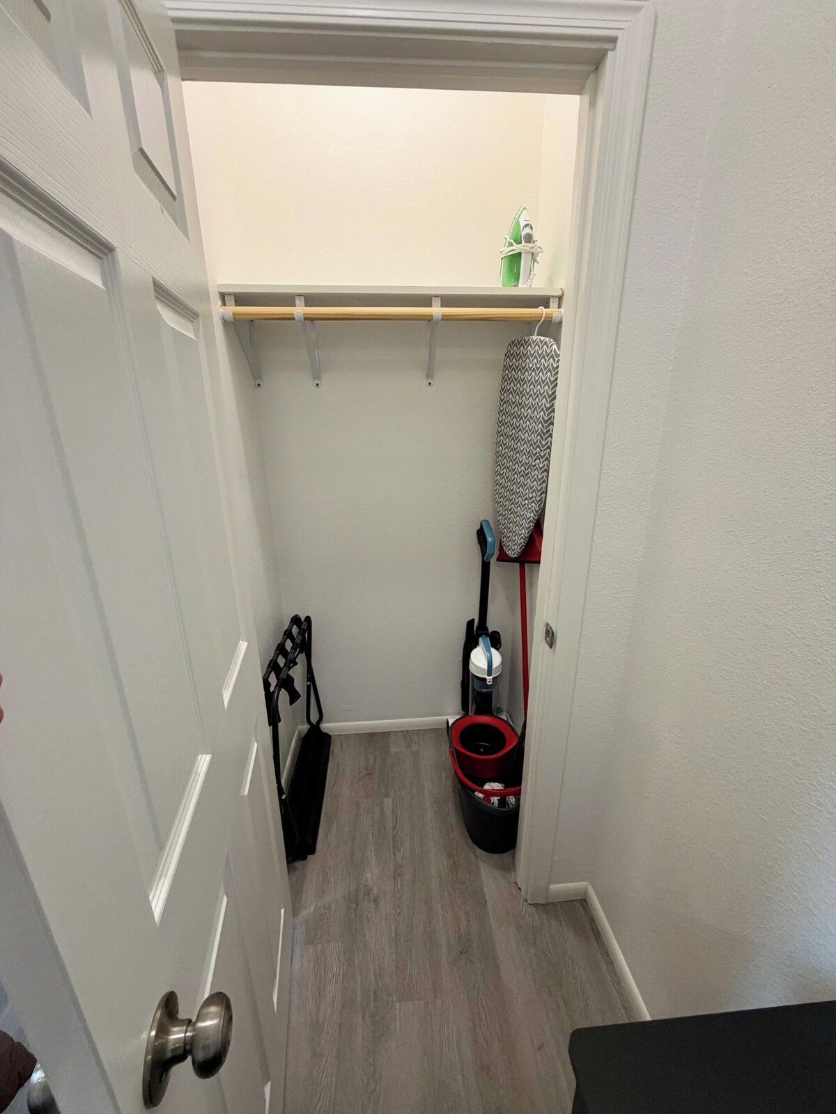 A narrow hallway with a wooden shelf, a hanging iron, a vacuum cleaner, and a black bag.