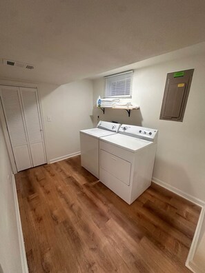 Un lavadero con lavadora y secadora, suelo de madera, una ventana con persianas y una puerta con paneles verticales.
