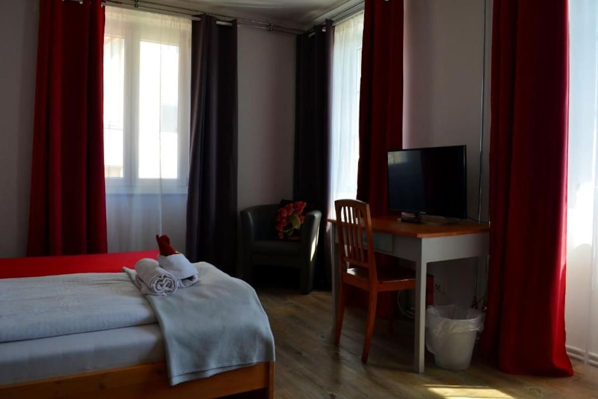 Habitación de hotel con cama, escritorio con televisor, una silla y ventana con cortinas.