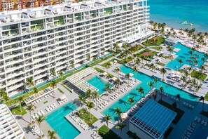 Ein mehrstöckiges Hotel mit einem großen Poolbereich, inklusive separater Schwimm- und Liegebereiche, umgeben von Palmen und Liegestühlen.