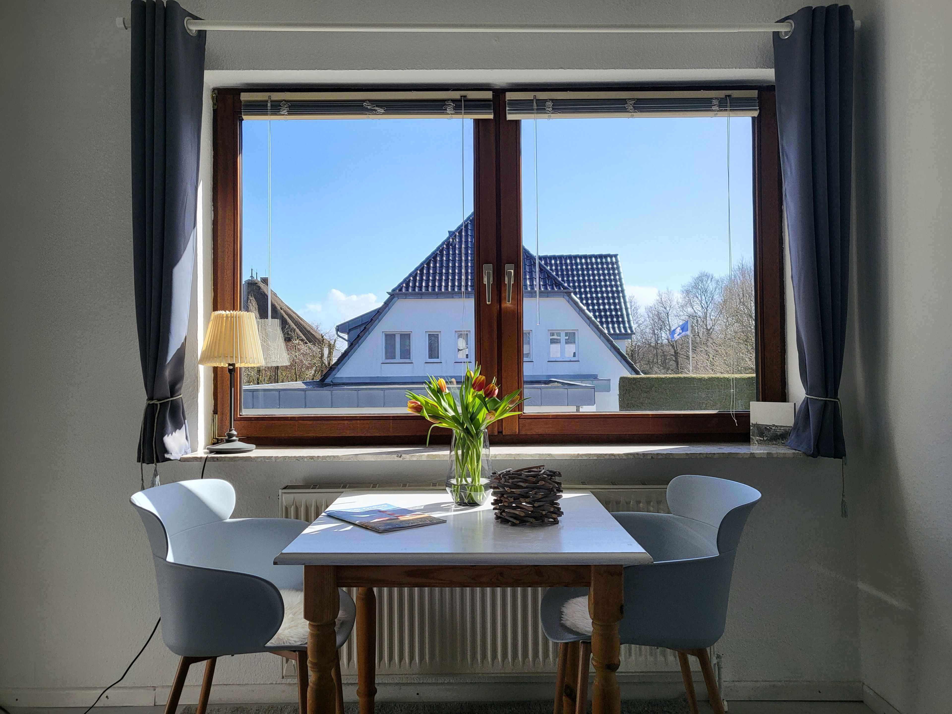 A room with a table and two chairs, a window with curtains, a radiator, and a view of a house outside.