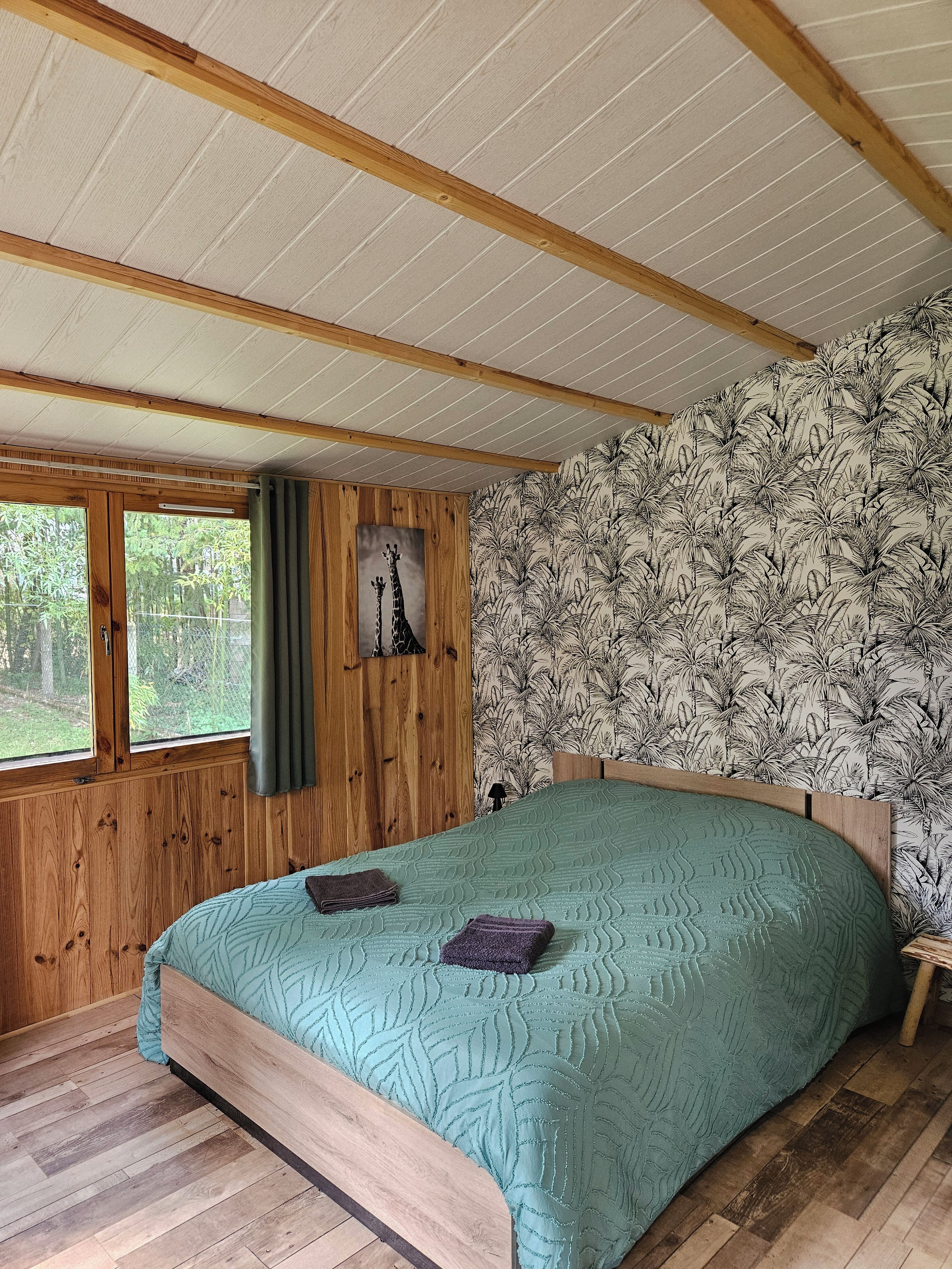 Een slaapkamer met een houten plafond, een bed met een bedovertrek met een patroon, een raam met gordijnen, en een wand met behang met een tropisch bladmotief.
