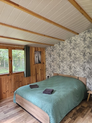 A bedroom with a wooden ceiling, a bed with a patterned bedspread, a window with curtains, and a wall with a tropical leaf wallpaper. - New Listing (Gond-Pontouvre)