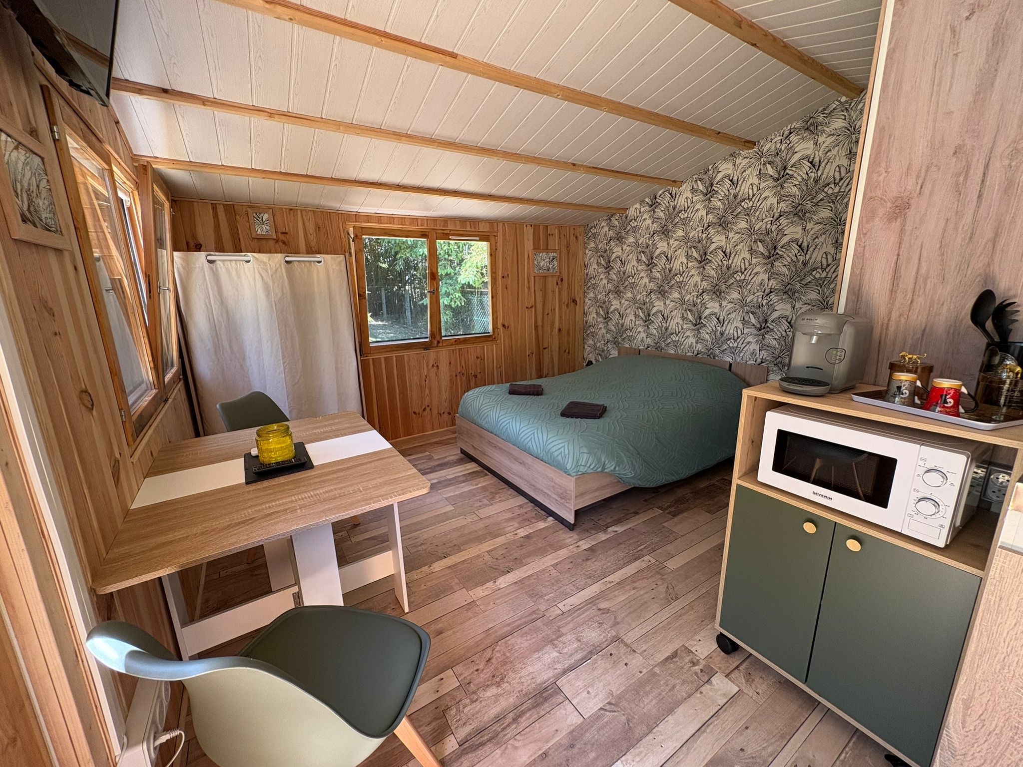 Een kleine kamer met een bed, een tafeltje, een stoel, een kitchenette met een magnetron en een raam met gordijnen.