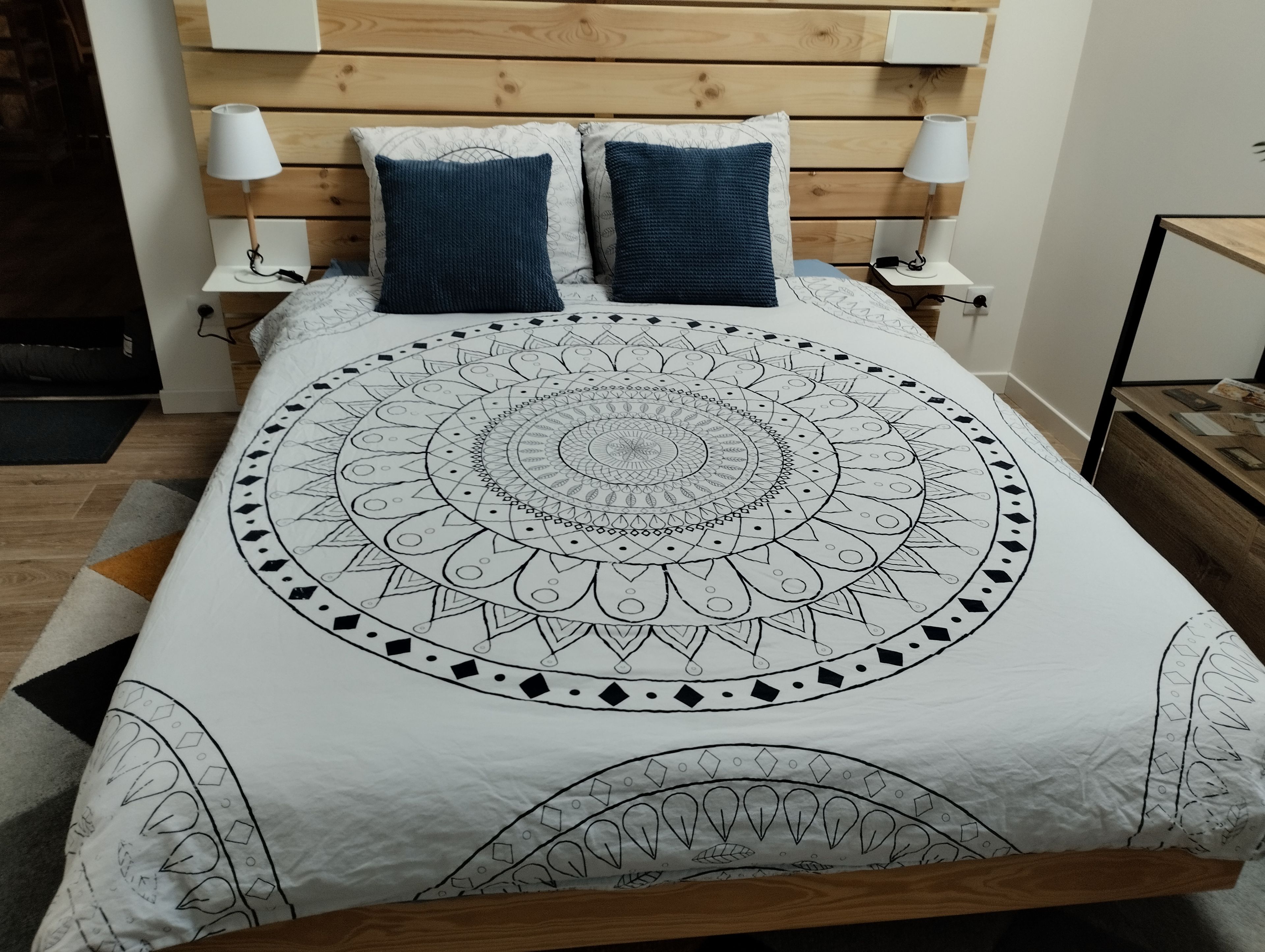 Ein Bett mit einem bettwandartigen, kreisförmigen Muster, zwei Kissen, ein Holz-Kopfteil, Nachttische mit Lampen und ein Nachtschränkchen.