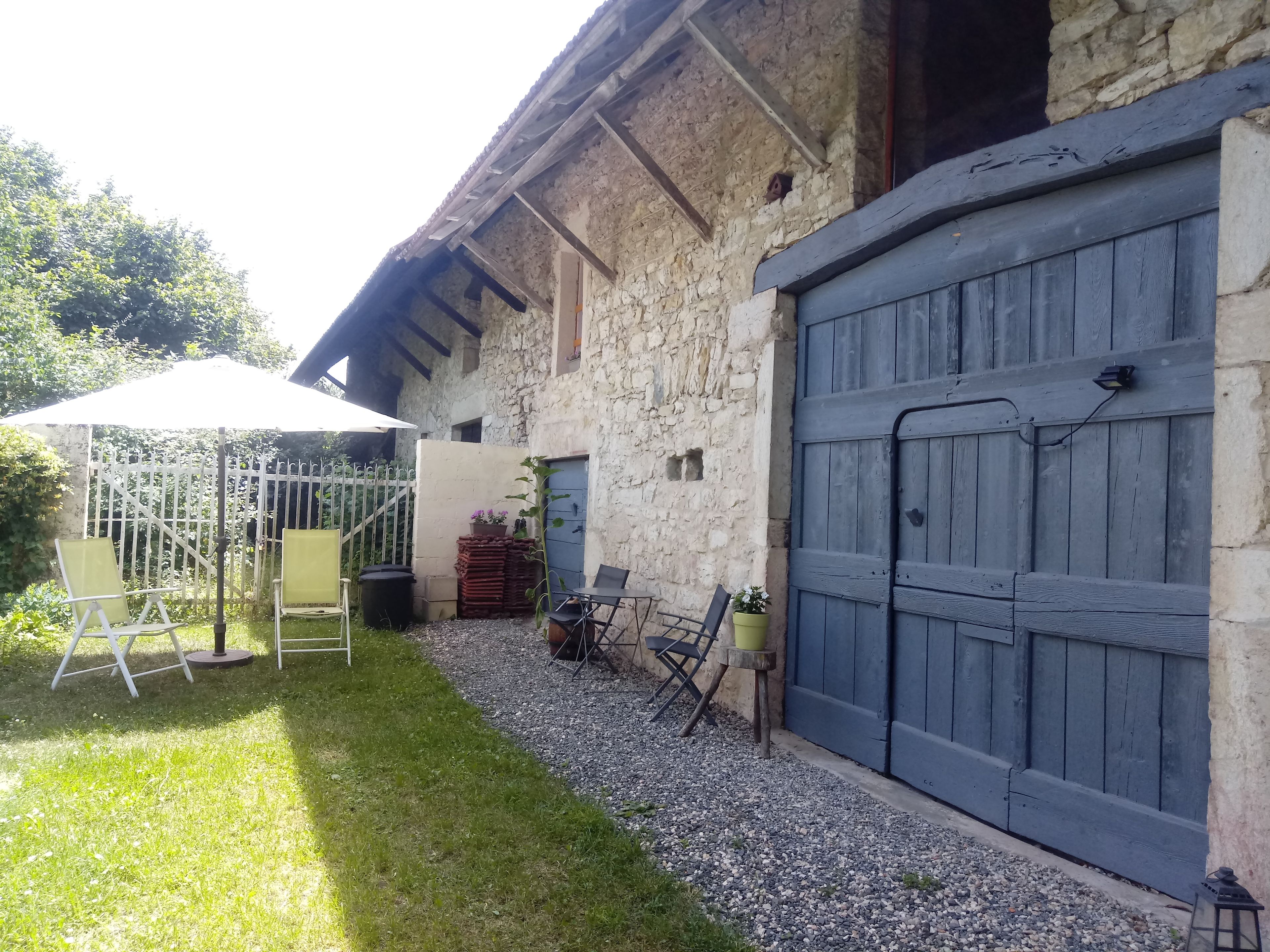 Ein Steinhaus mit einer großen Holztür zum Carport, einer überdachten Terrasse mit Stühlen und Tisch sowie einem Kiesweg.