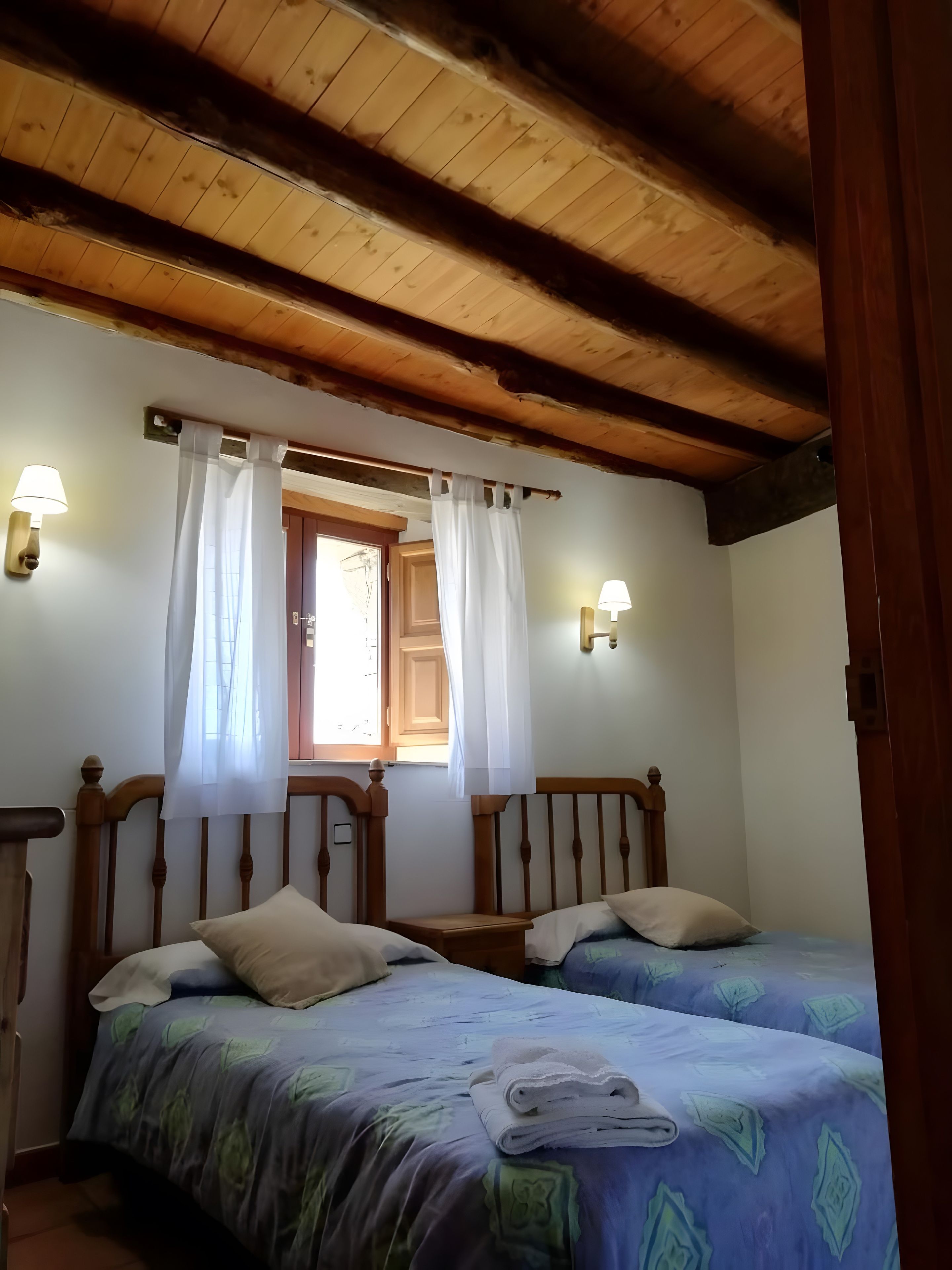 A room with two beds, wooden beams on the ceiling, and two wall-mounted lamps.