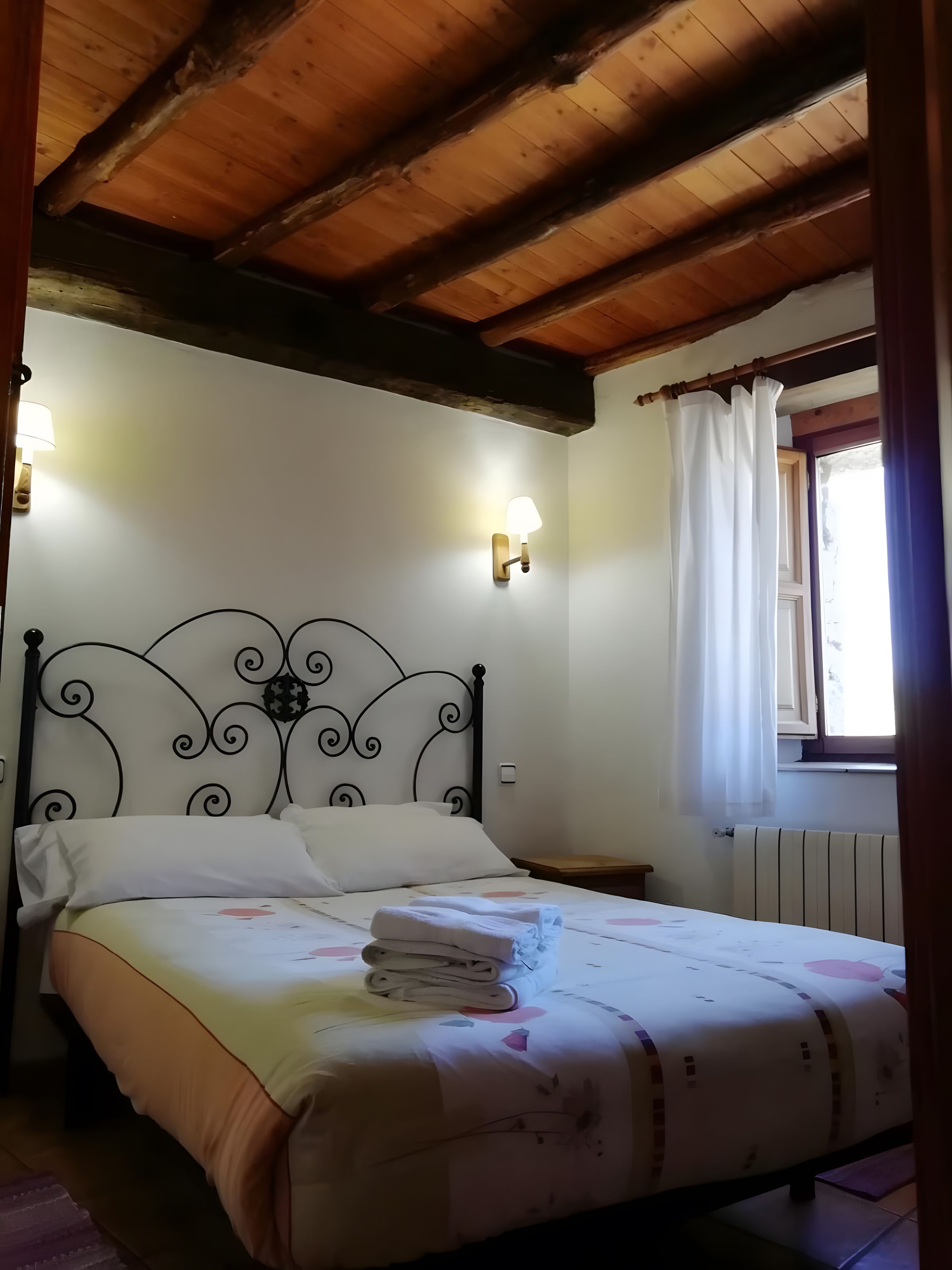 A bedroom with a bed, a nightstand, a wall-mounted lamp, and a window with curtains.