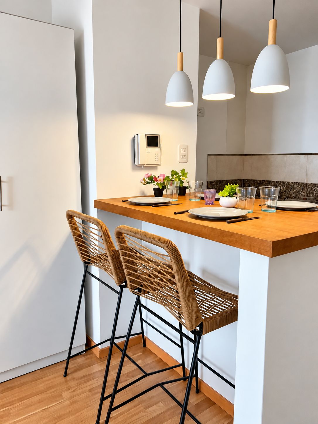 Una cocina moderna con una isla de madera, dos sillas de mimbre, lámparas colgantes y una pequeña planta en maceta.