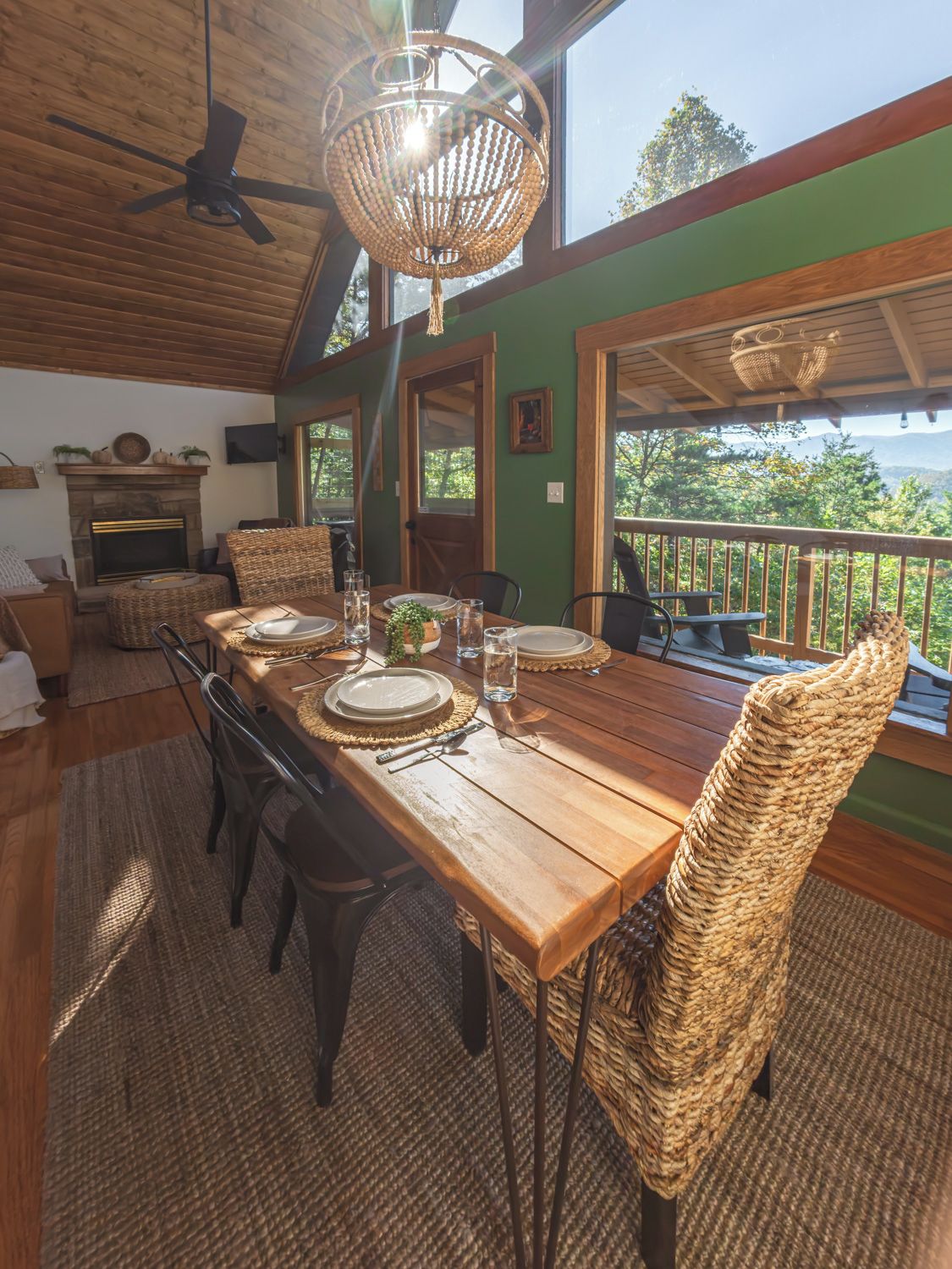 A dining area with a wooden table set for four, surrounded by wicker chairs, a fireplace, and a view of the outdoors.