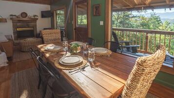 A dining area with a wooden table set for four, surrounded by wicker chairs, a fireplace, and a view of the outdoors.