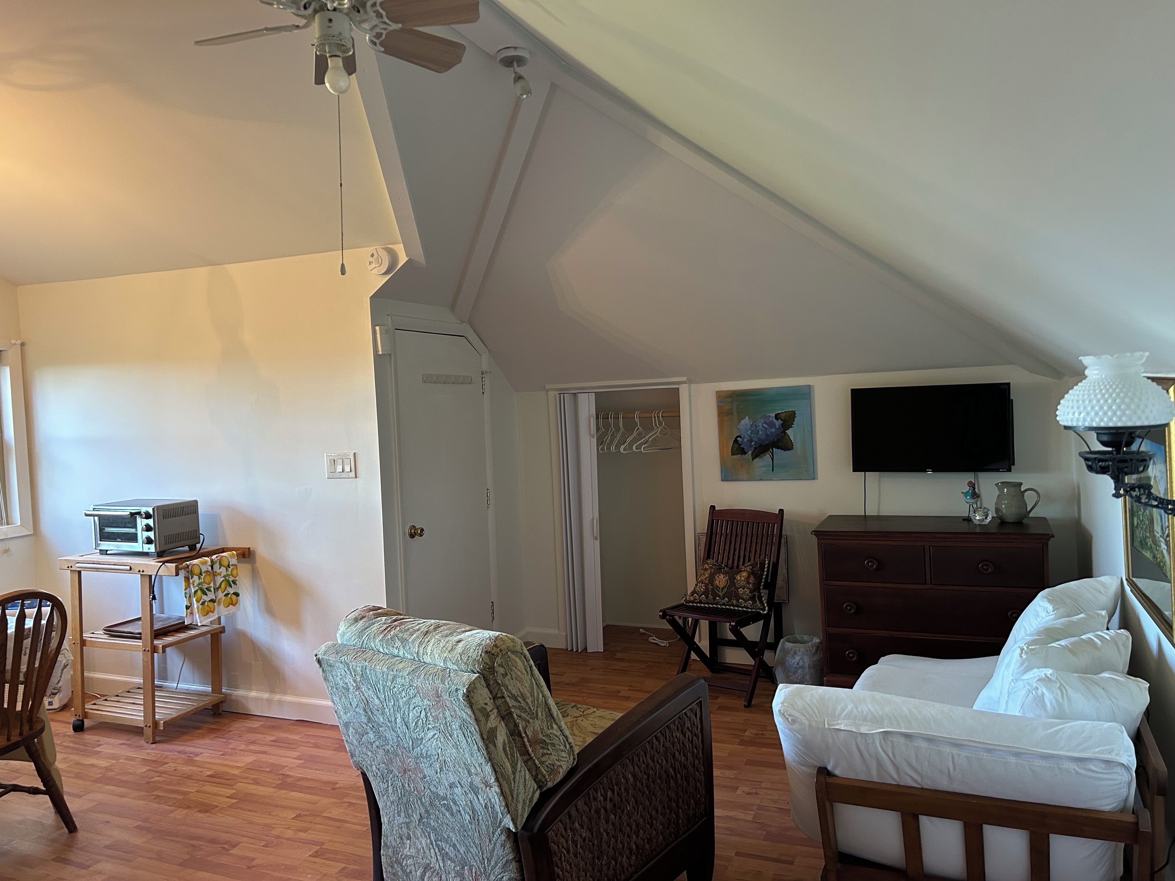 Una habitación con ventilador de techo, televisión, cómoda de madera, cama con ropa blanca y una silla con funda estampada.