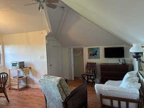 Ein Zimmer mit Deckenventilator, Fernseher, Holzkommode, Bett mit weißer Bettwäsche und Sessel mit gemustertem Bezug.
