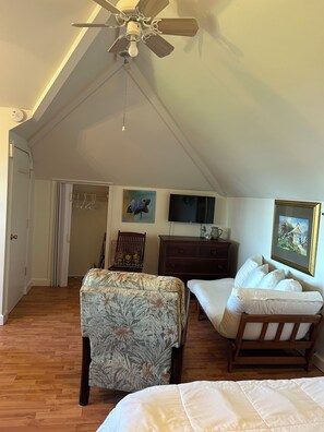 Ein Schlafzimmer mit geneigter Decke, Deckenventilator, Bett, Sessel, Kommode und Fernseher.