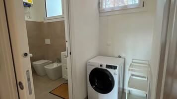 A compact laundry room with a washing machine, a drying rack, and a window with a view of trees.