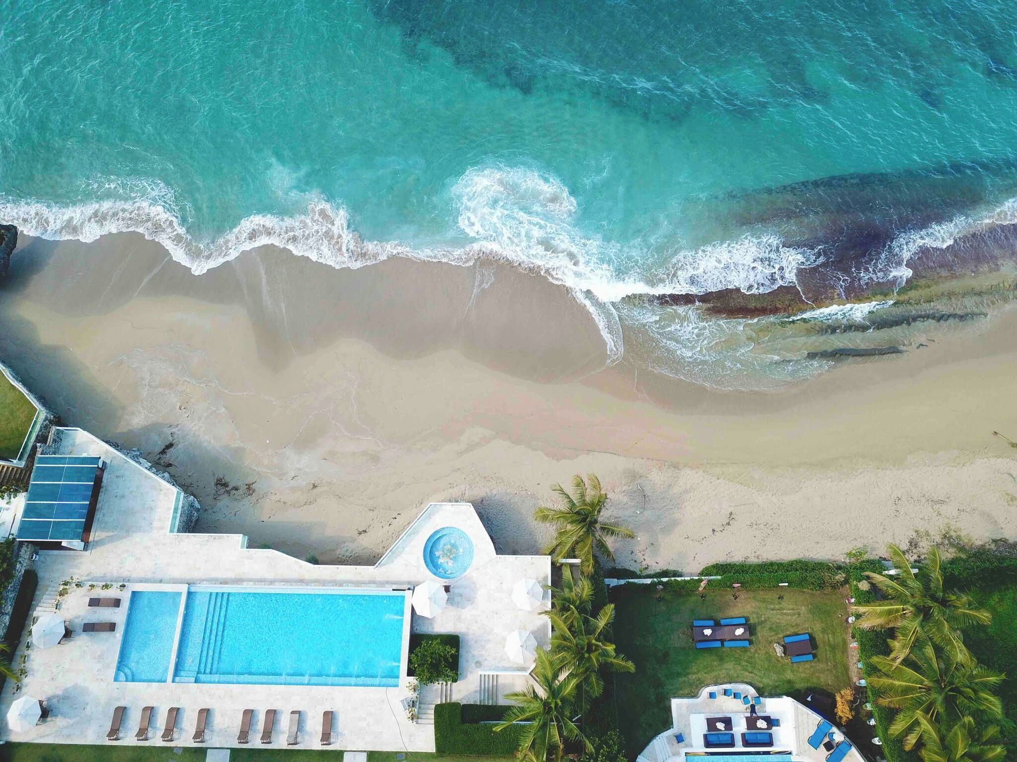 Vista aérea de una propiedad frente a la playa con piscina, sillas de descanso y vista al océano.
