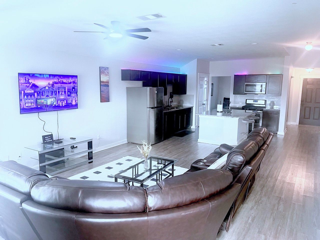 A modern living room with a leather sofa, a flat-screen TV, a glass coffee table, and a kitchen area in the background.