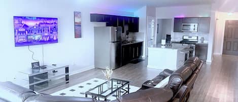 A modern living room with a leather sofa, a flat-screen TV, a glass coffee table, and a kitchen area in the background.