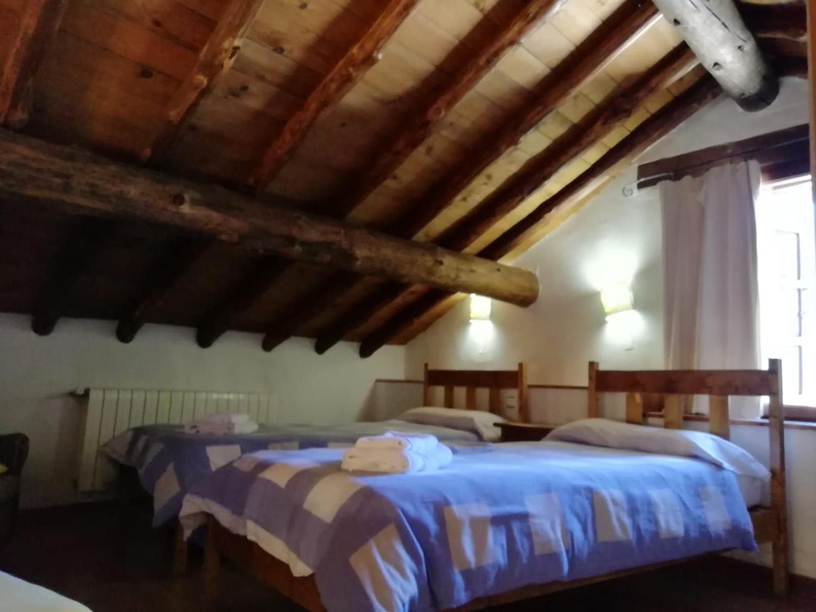 Een kamer met een schuin houten plafond, een bed met een blauw-witte sprei, twee nachtkastjes en een raam met gordijnen.