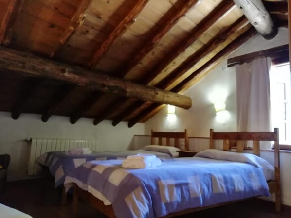 Een kamer met een schuin houten plafond, een bed met een blauw-witte sprei, twee nachtkastjes en een raam met gordijnen.