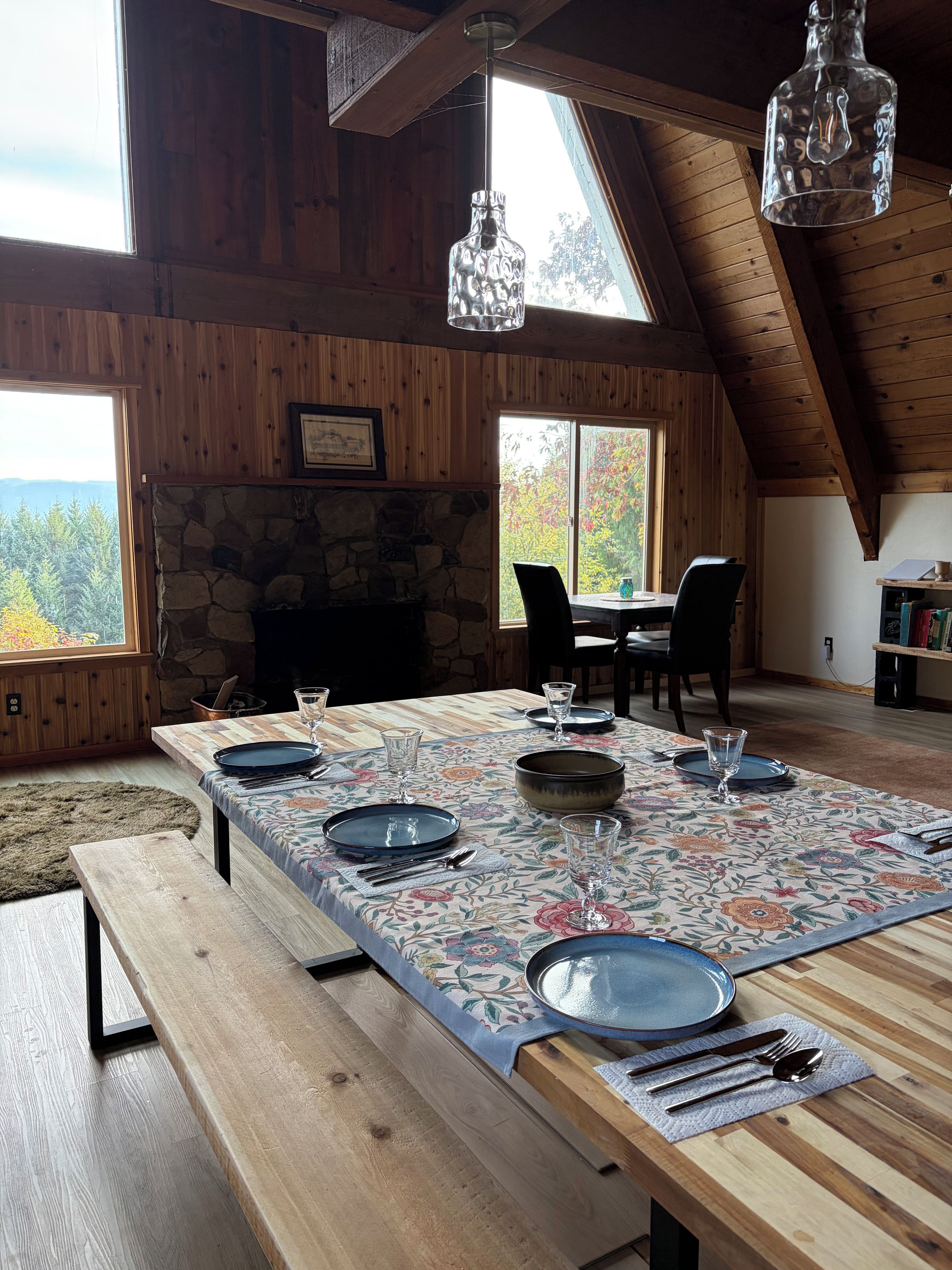 Un espace repas avec une longue table dressée pour six personnes, des poutres en bois au plafond, une cheminée en pierre et une vue sur les arbres par les fenêtres.