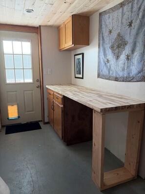 Une cuisine avec des meubles de rangement en bois, un plan de travail, une porte avec un judas et un mur orné d’un tableau encadré.
