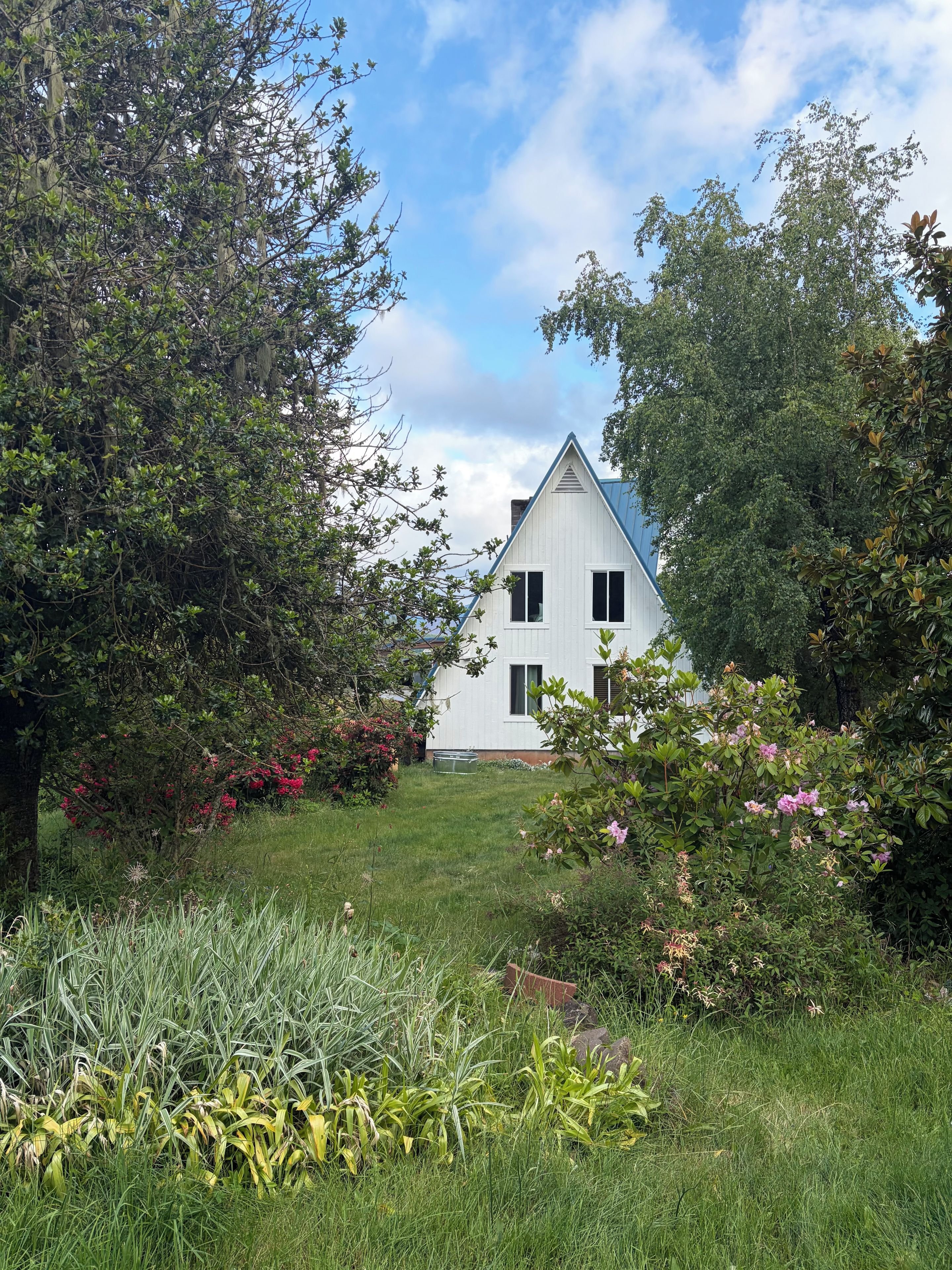 Une maison blanche avec un toit triangulaire, entourée d’une végétation luxuriante et de plantes en fleurs.