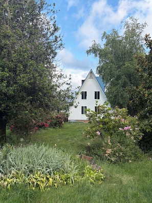 Une maison blanche avec un toit triangulaire, entourée d’une végétation luxuriante et de plantes en fleurs.